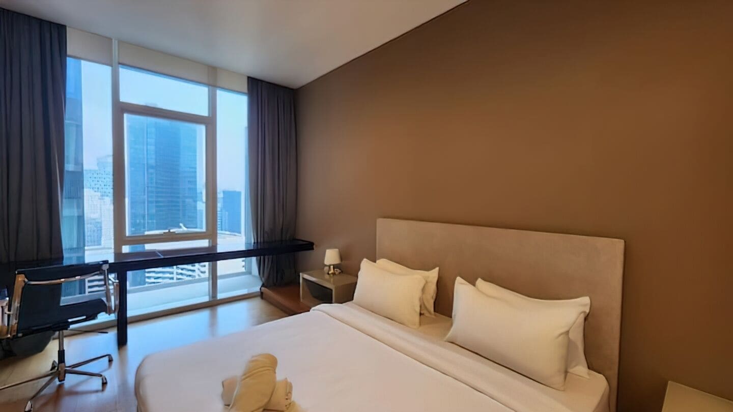 A hotel room with a large bed, a desk, and a view of a cityscape through large windows.