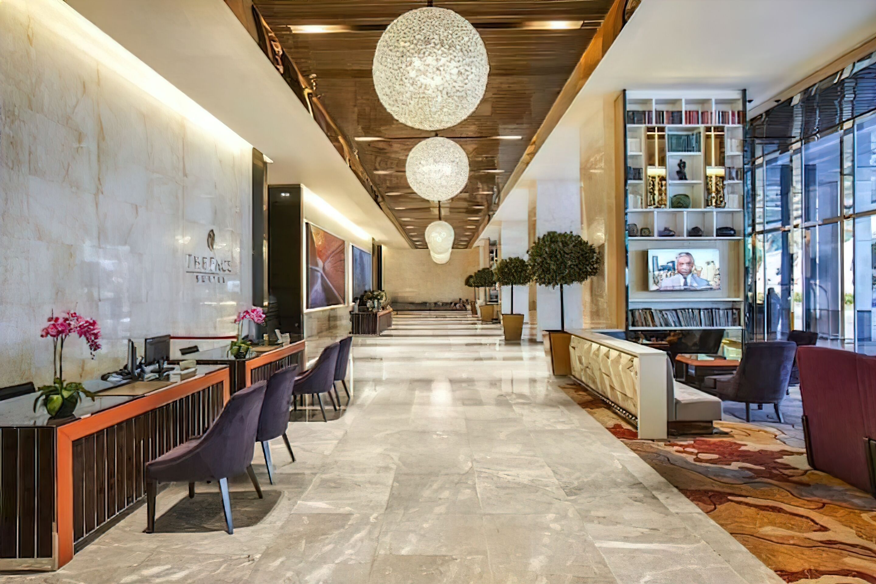 A hotel reception area with a marble reception desk, purple chairs, a large chandelier, and a bookshelf with books and decorative items.