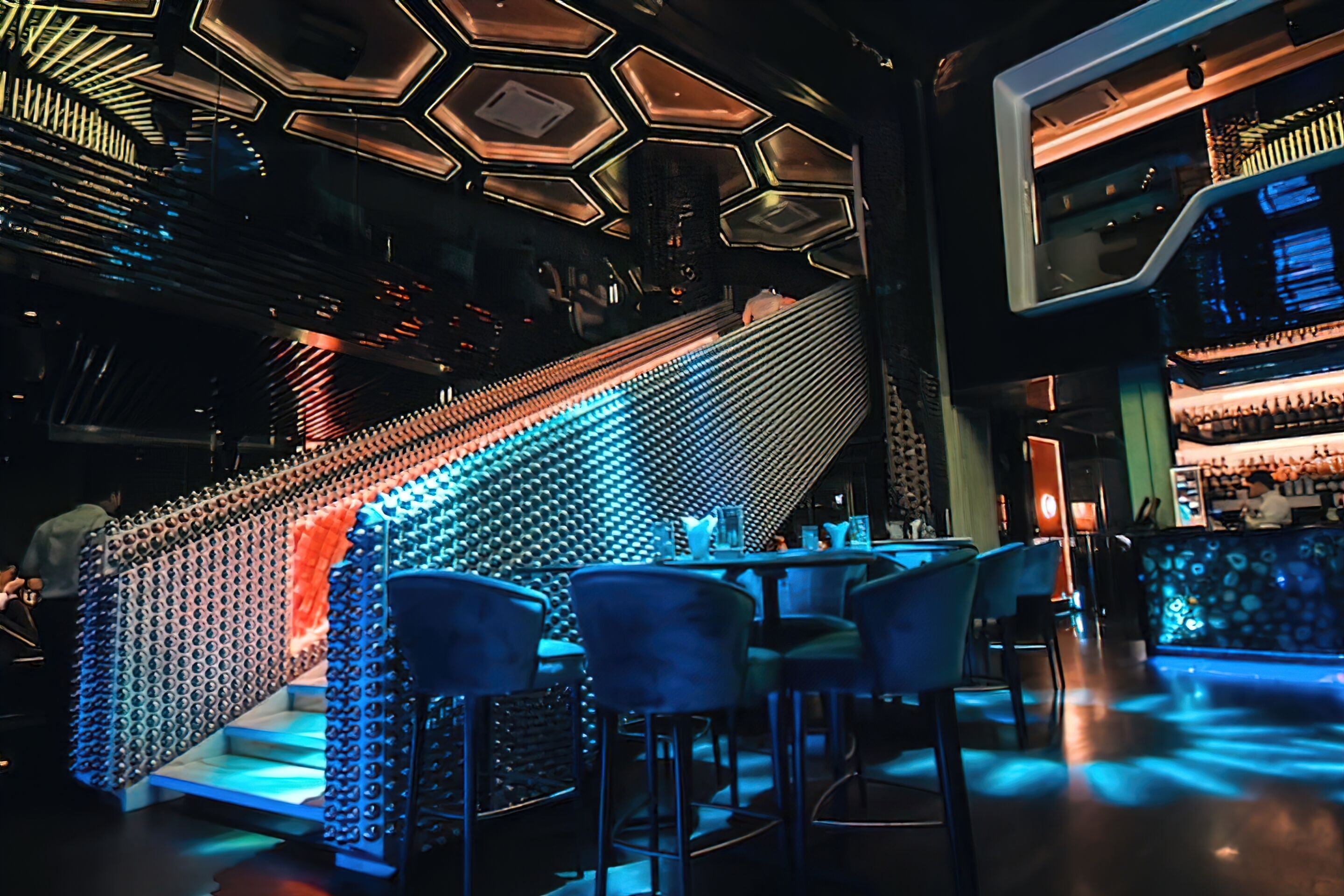 A modern bar area with a unique geometric light design on the ceiling, a staircase with illuminated steps, and bar seating.