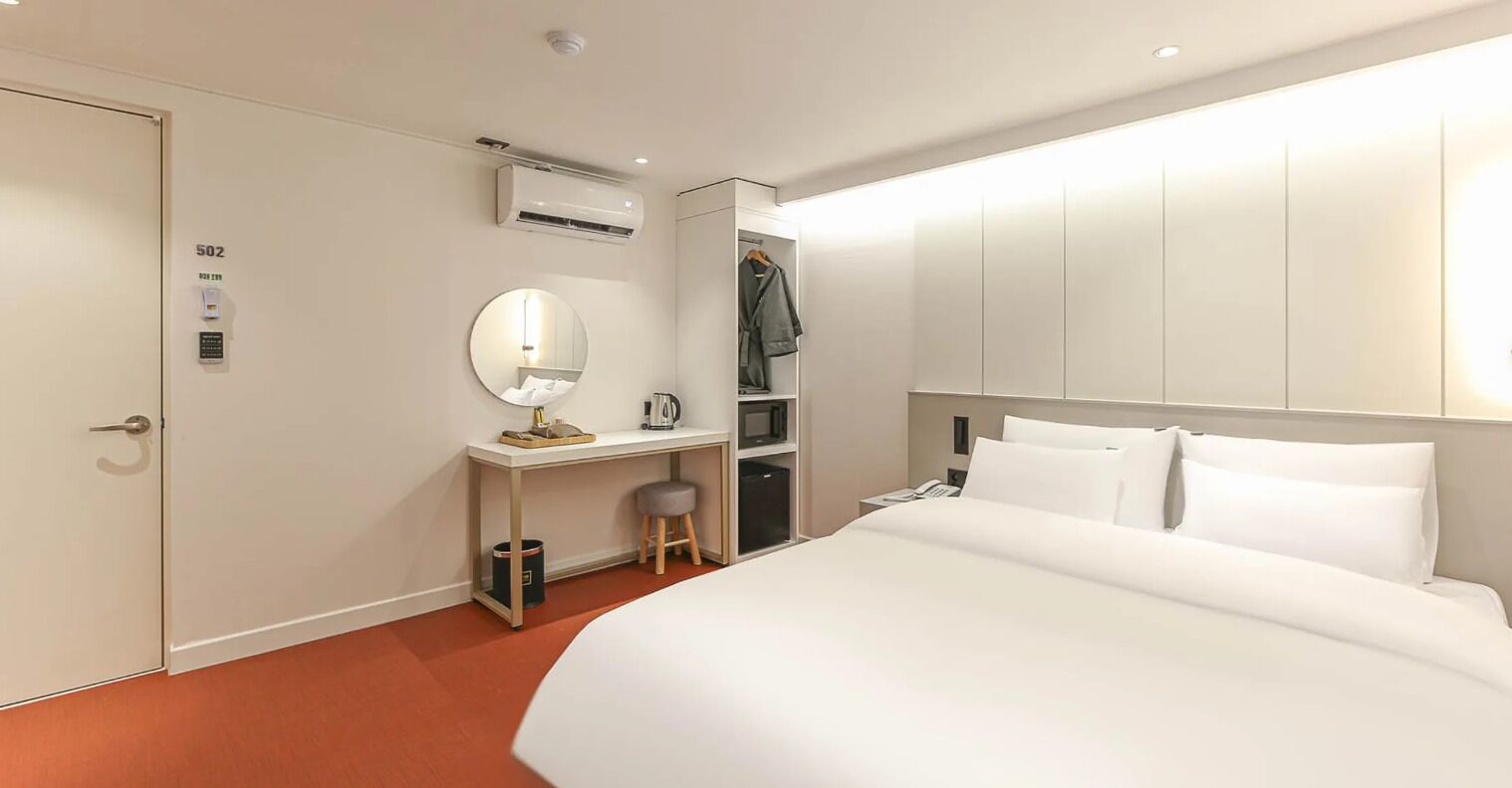 A hotel room with a bed, a desk with a chair, a mirror, an air conditioning unit, and a wardrobe.