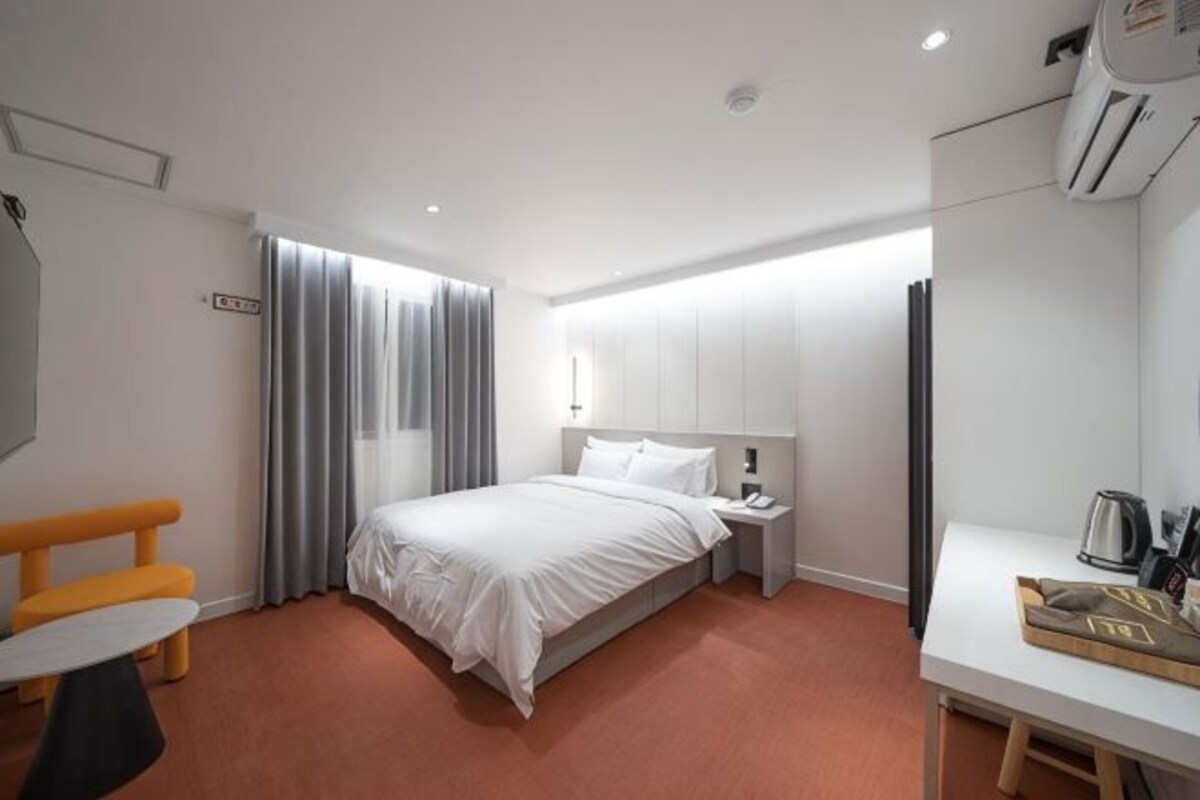A hotel room with a bed, bedside table, chair, and a desk with a kettle.