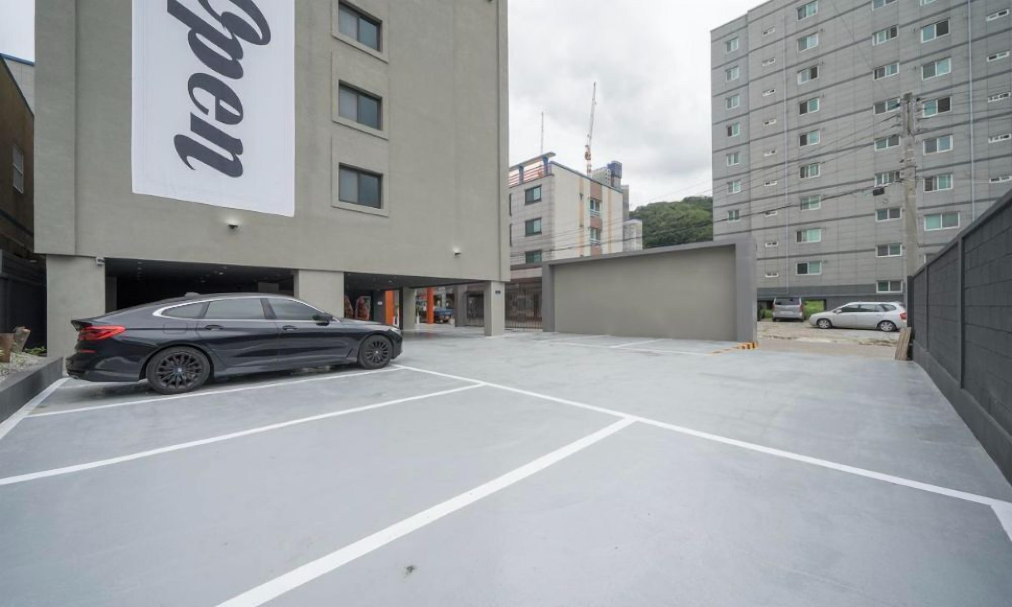A parking area with a black car, a building with a large sign, and a multi-story residential building in the background.