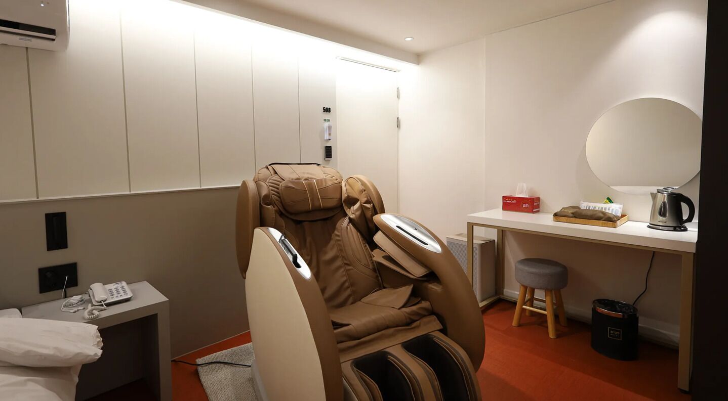 A massage chair, a desk with a mirror, a telephone, and a small stool in a room with a red carpet.