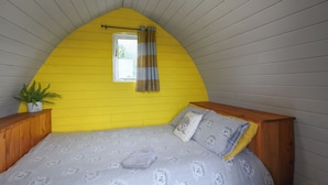 A bedroom with a sloped ceiling, a bed with patterned bedding, a window with curtains, and a potted plant. - The Hedgehog Pod - UK50067 (Welshpool)