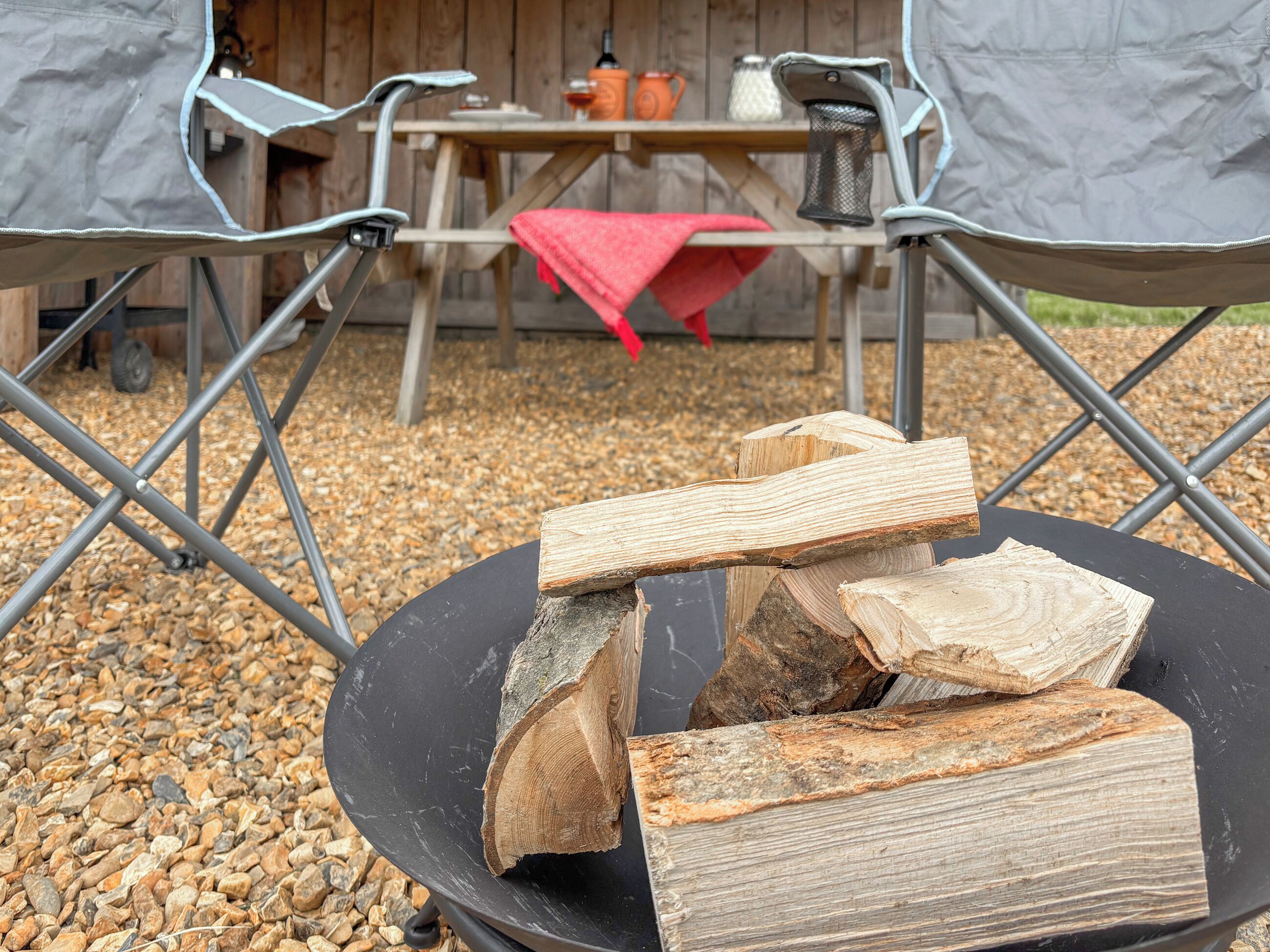 Una fogata con leña en un recipiente negro, rodeada de sillas de camping y una mesa con un mantel rojo.