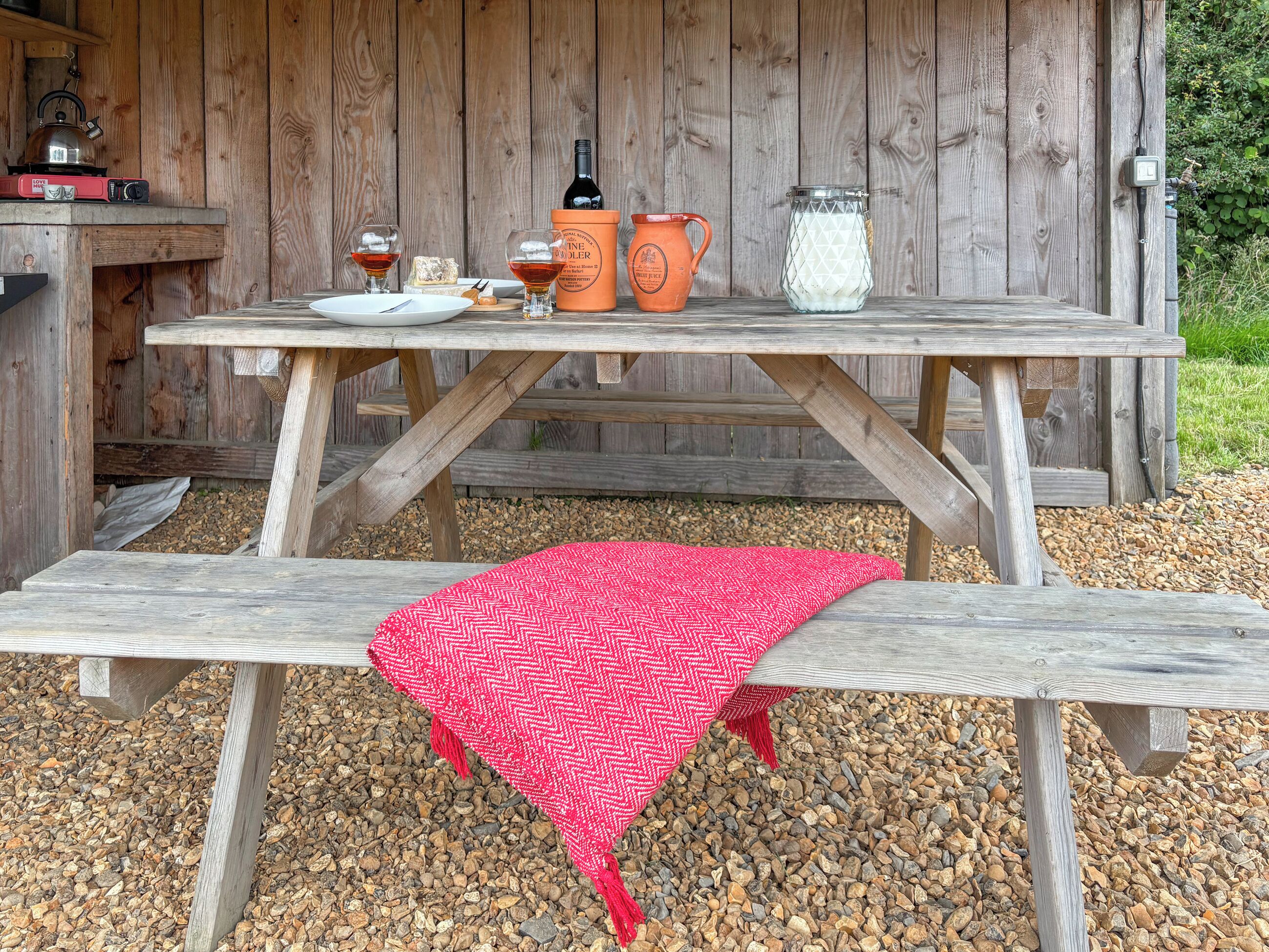 A wooden picnic table with a red blanket, a bottle of wine, glasses, and a jug of milk.