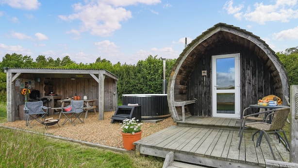 A rustic outdoor setting with a wooden cabin featuring a hot tub, a barbecue area, and a small shed with a table and chairs. - The Hedgehog Pod - UK50067 (Welshpool)
