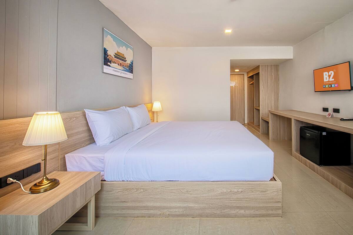 A hotel room with a bed, bedside table, lamp, TV, and a small kitchenette area.