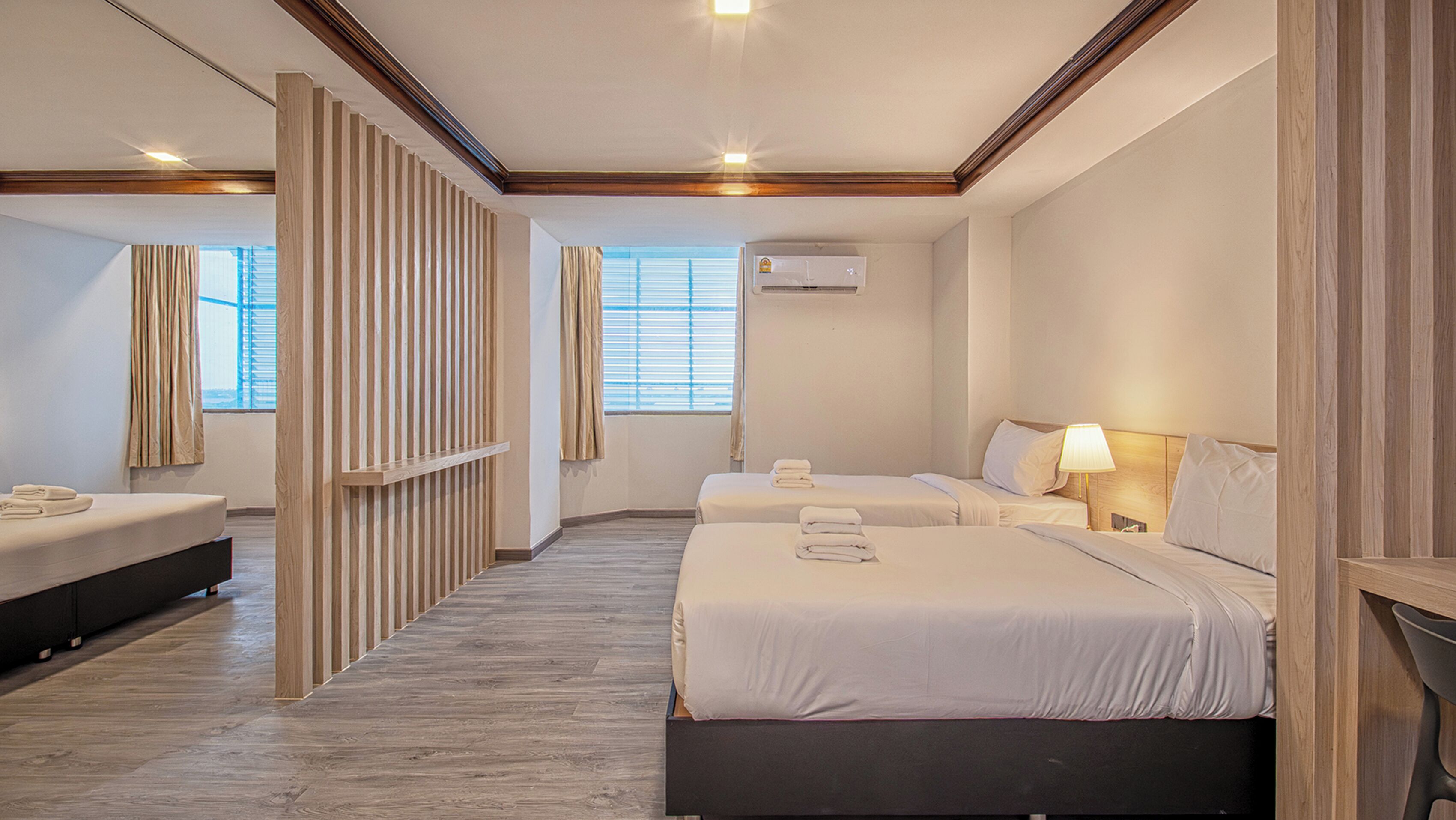 A hotel room with two beds, a wooden partition, a window with blinds, an air conditioning unit, and a bedside lamp.