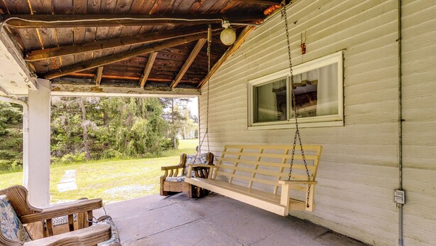 A covered porch with a wooden bench and chairs, a hanging swing, and a view of a forested area. - Pet-Friendly Smithmill Retreat w/ Yard! (Smithmill)