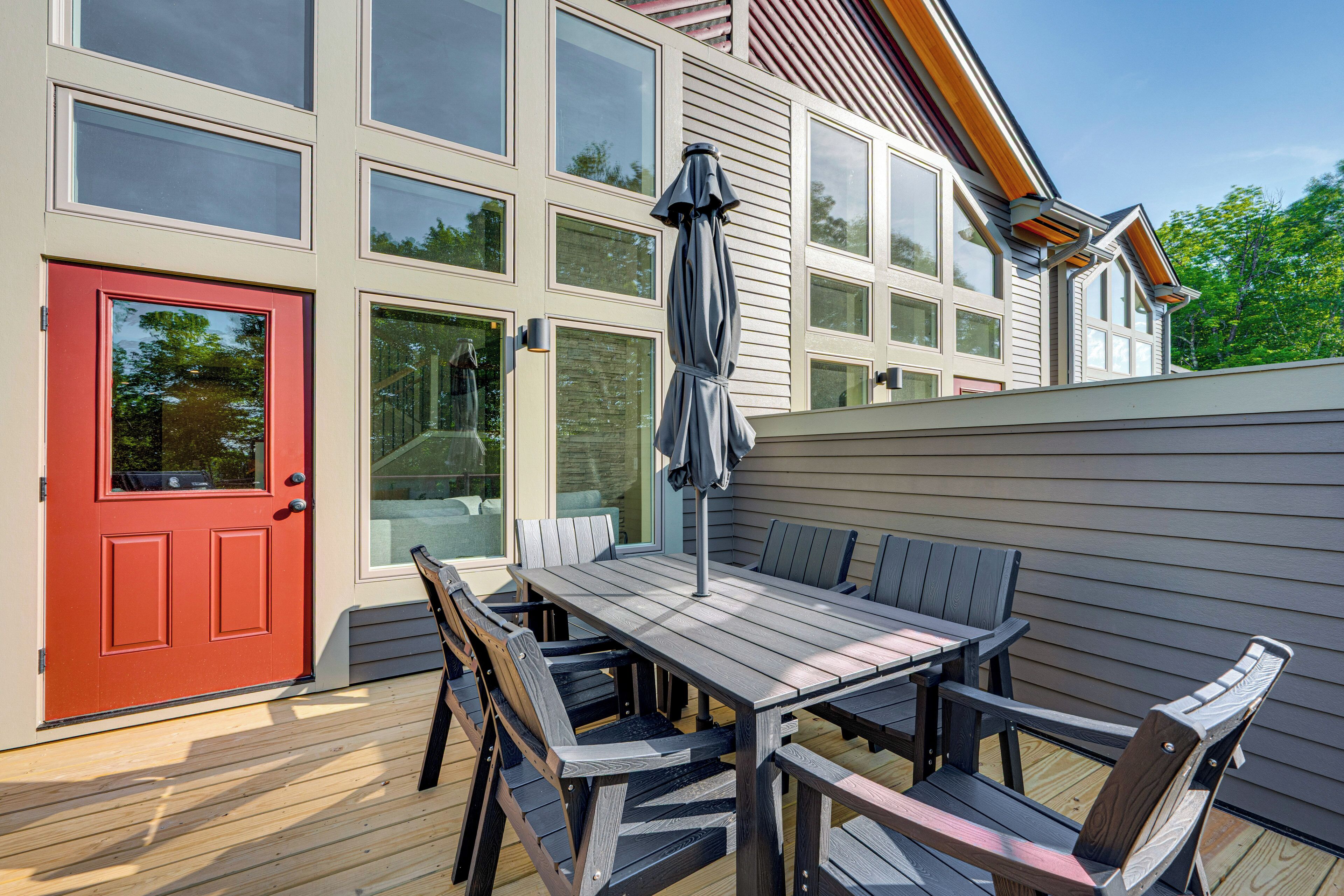 Une terrasse avec une table et des chaises en bois, une porte rouge et de grandes fenêtres.