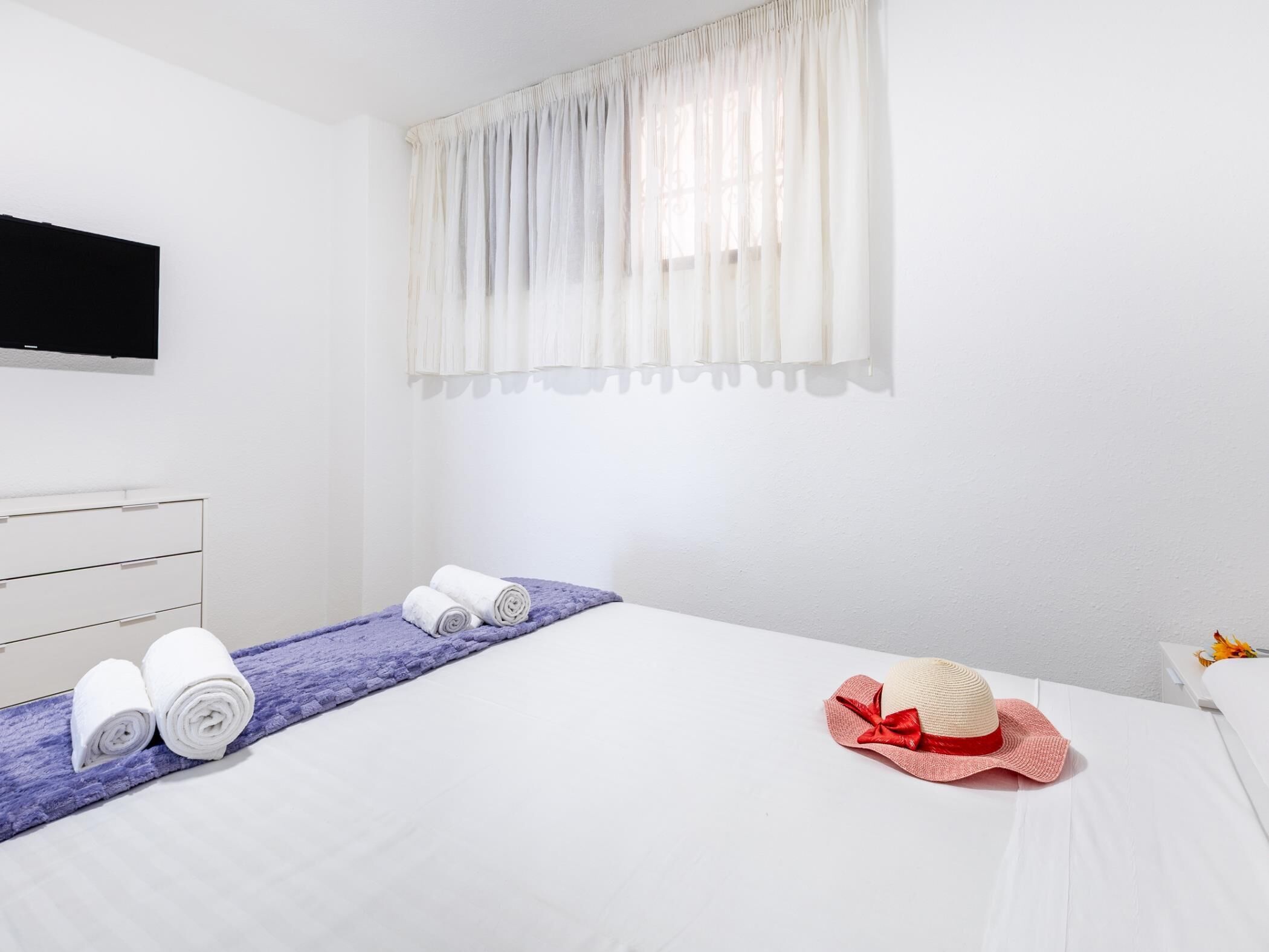 A hotel room with a bed, a television, a window with curtains, a white dresser, and a rolled towel on the bed.