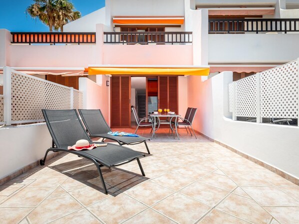Eine sonnige Terrasse mit zwei Liegestühlen, einem Tisch mit Stühlen und einem überdachten Bereich.