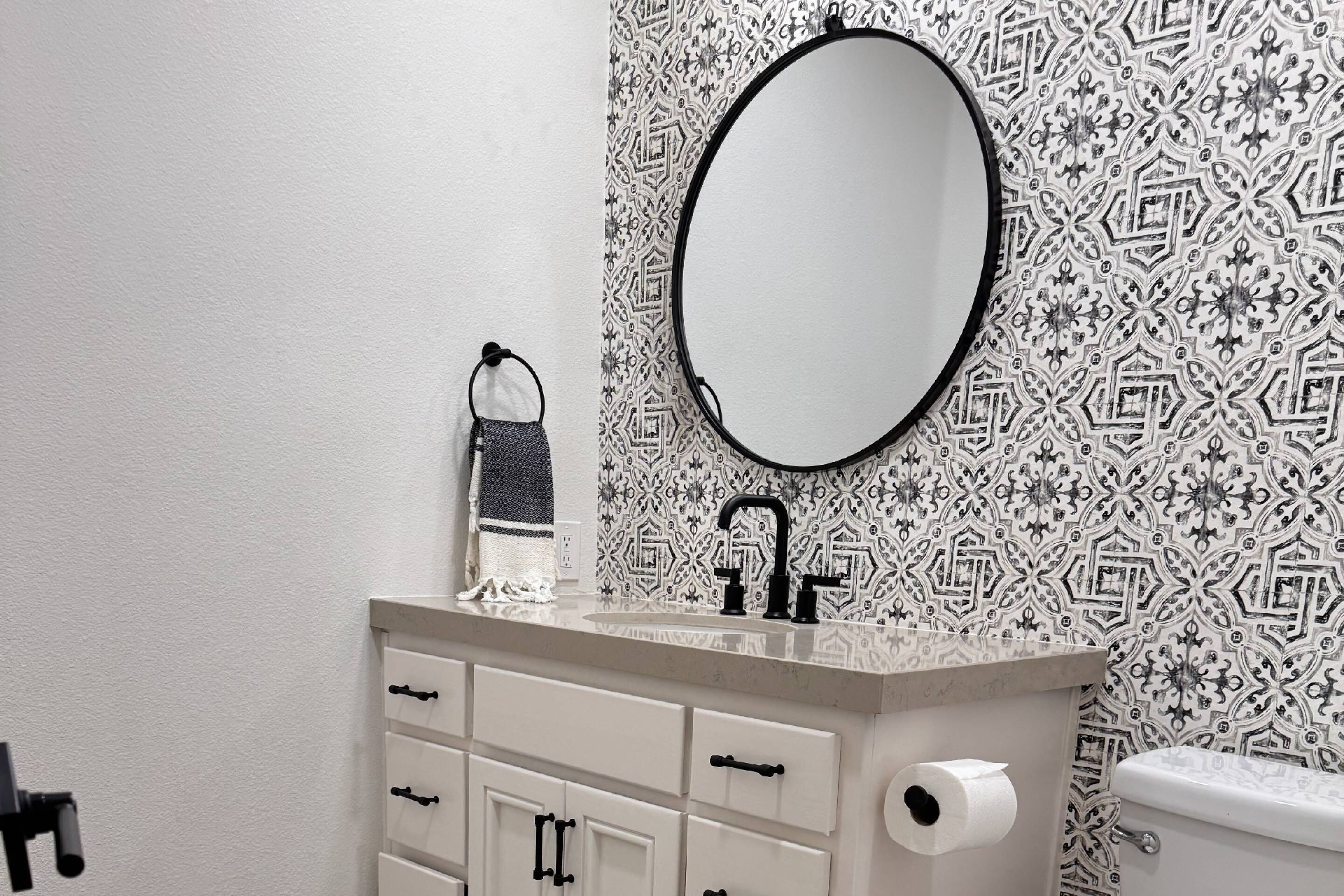 Een badkamer met behang met een patroon, een ronde spiegel, een wastafel met een kraan, een handdoekhouder en een toilet.