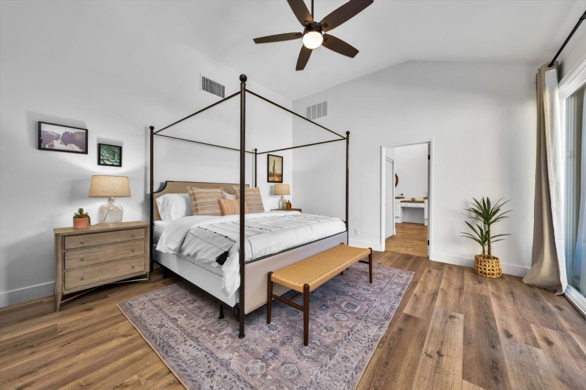 A four-poster bed with a wooden frame, a bench at the foot, a nightstand with a lamp, and a large window with curtains.