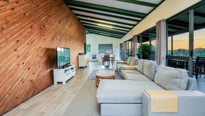 Ein modernes Wohnzimmer mit einer Wand in Holzoptik, einer grauen Sofagarnitur, einem Fernseher und einem Essbereich.