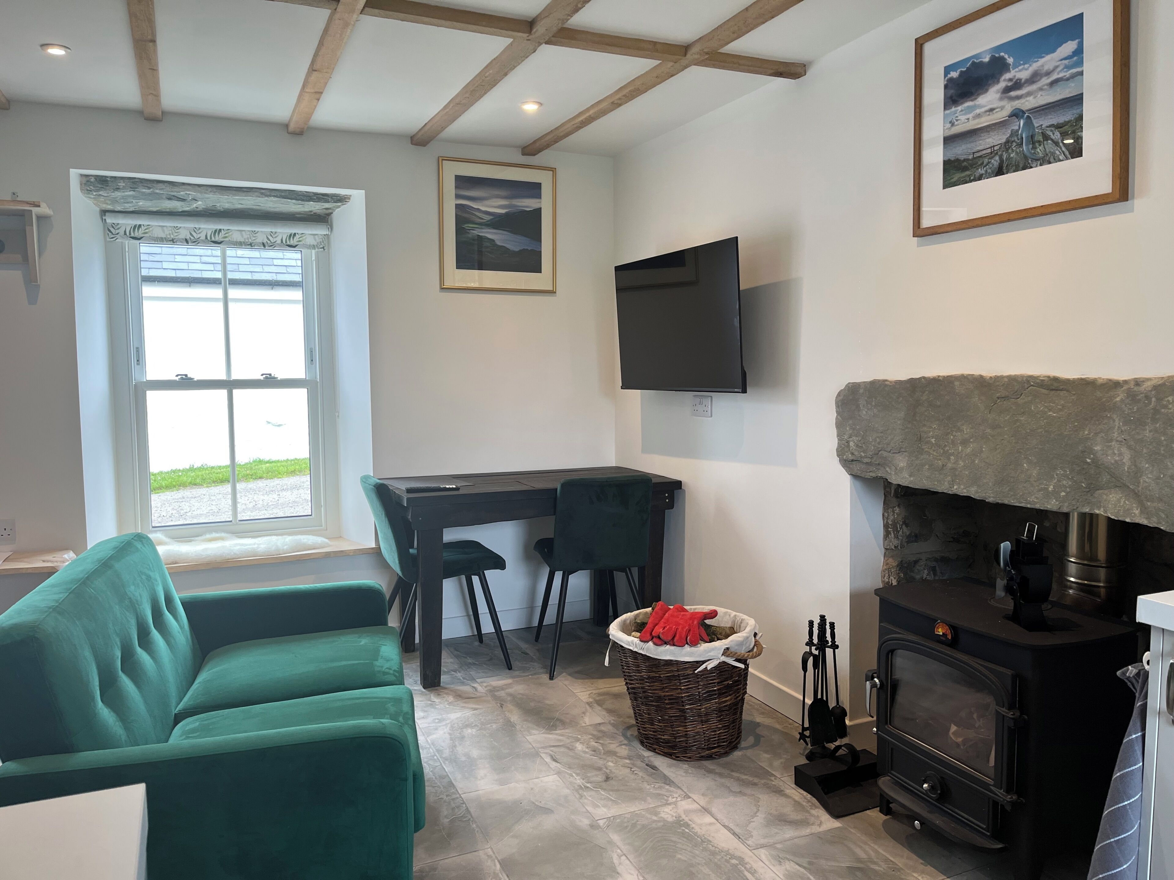 A room with a stone fireplace, a television mounted on the wall, a desk with chairs, and a large window with a view of the outdoors.