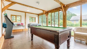 Una mesa de billar en una habitación con vigas de madera, ventanas amplias y vistas a la vegetación exterior.