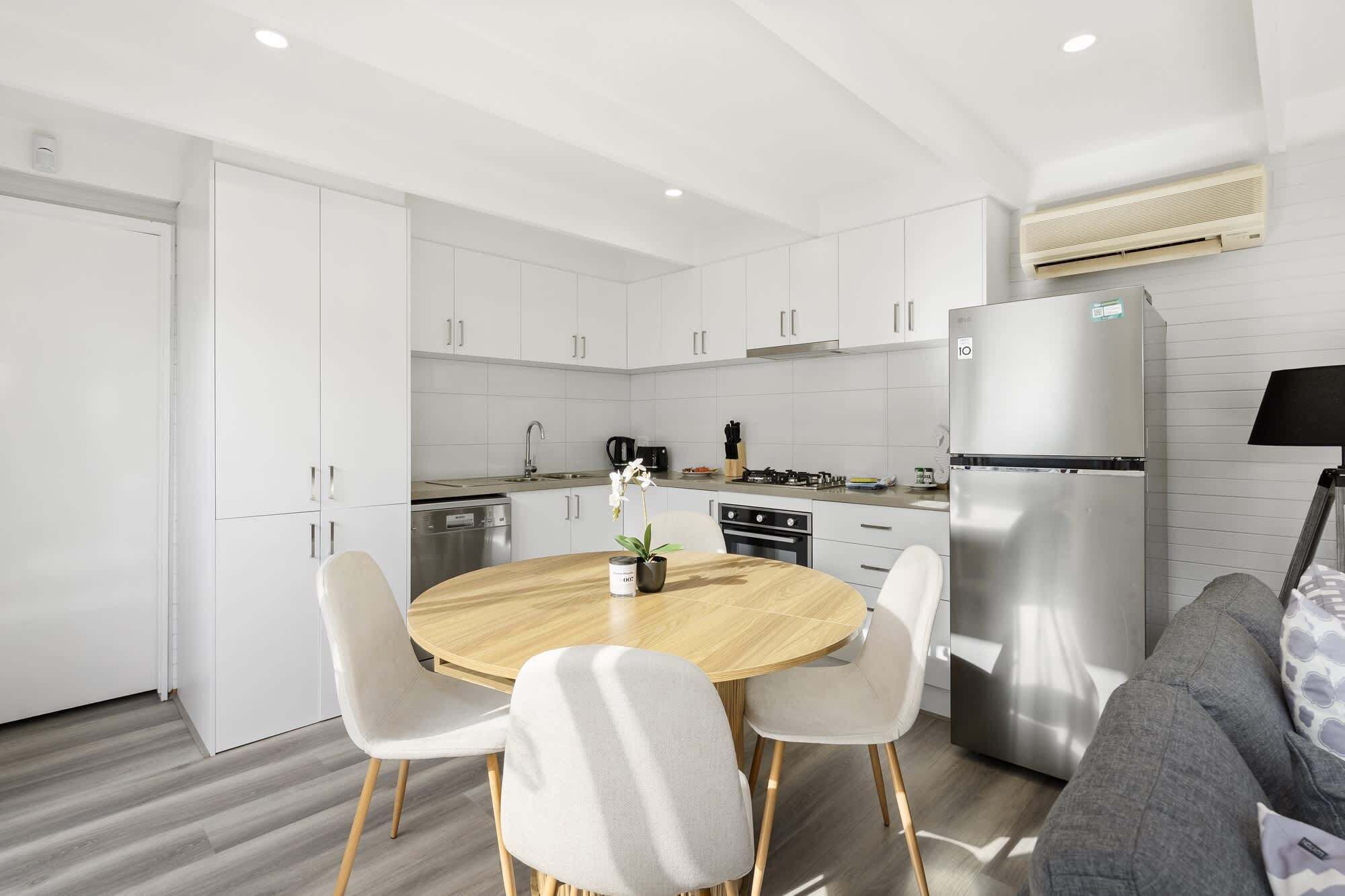 A modern kitchen with white cabinetry, a stainless steel refrigerator, and a round wooden dining table with four chairs.