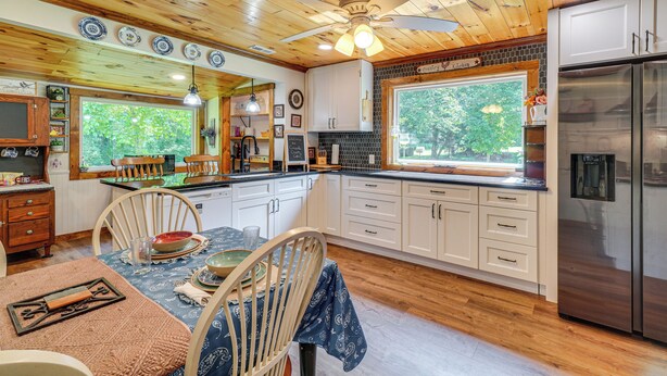 Een keuken met houten plafond, witte kasten, een roestvrijstalen koelkast, en een eethoek met tafel en stoelen.
