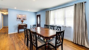 A dining room with a wooden table and chairs, a painting on the wall, and a window with curtains.