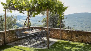 An outdoor dining area with a table and chairs, surrounded by stone walls and a pergola with vines.