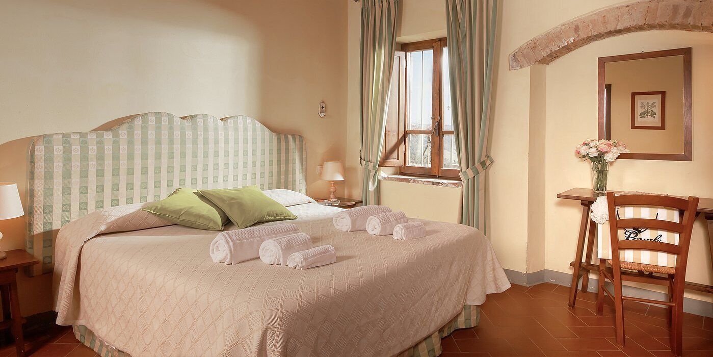 Een hotelkamer met een bed, groene kussens, een stoel, een tafel met een vaas en een raam met gordijnen.
