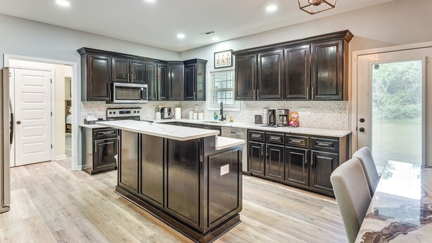 A modern kitchen with dark cabinetry, a central island, and a dining area with a marble table. - 11 Mi to Troy University: Family Home w/ Yard (Troy)