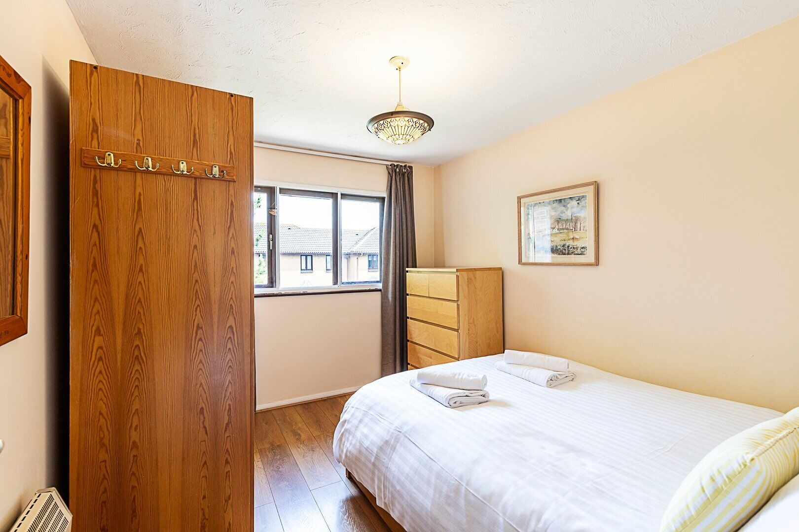 A bedroom with a wooden wardrobe, a bed with white linens, a dresser, a framed picture on the wall, and a window with curtains.