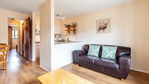 Ein modernes Wohnzimmer mit einer braunen Ledersofa, einem Holztisch und einem Essbereich mit Holztisch und Stühlen. Im Hintergrund ist eine Küche mit weißen Schränken und einer hängenden Kochtopfhalterung erkennbar.