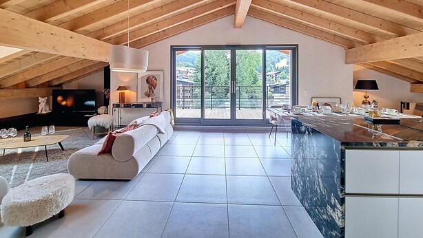 Un loft spacieux avec un plafond haut, une grande porte-fenêtre coulissante donnant sur un balcon, un coin salon confortable avec un canapé et un pouf, et un espace cuisine doté d’un comptoir en marbre.