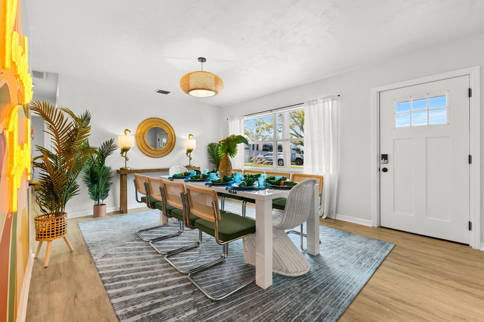 Un coin repas avec une table blanche dressée pour six personnes, entourée de chaises dotées de coussins verts. Des plantes en pot sont disposées sur la table et un grand miroir orne le mur.