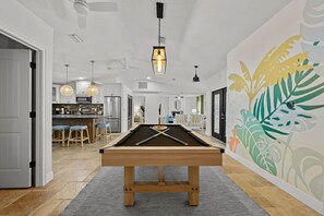 A pool table in a modern living room with a kitchen in the background, a dining area, and a wall with a tropical mural.