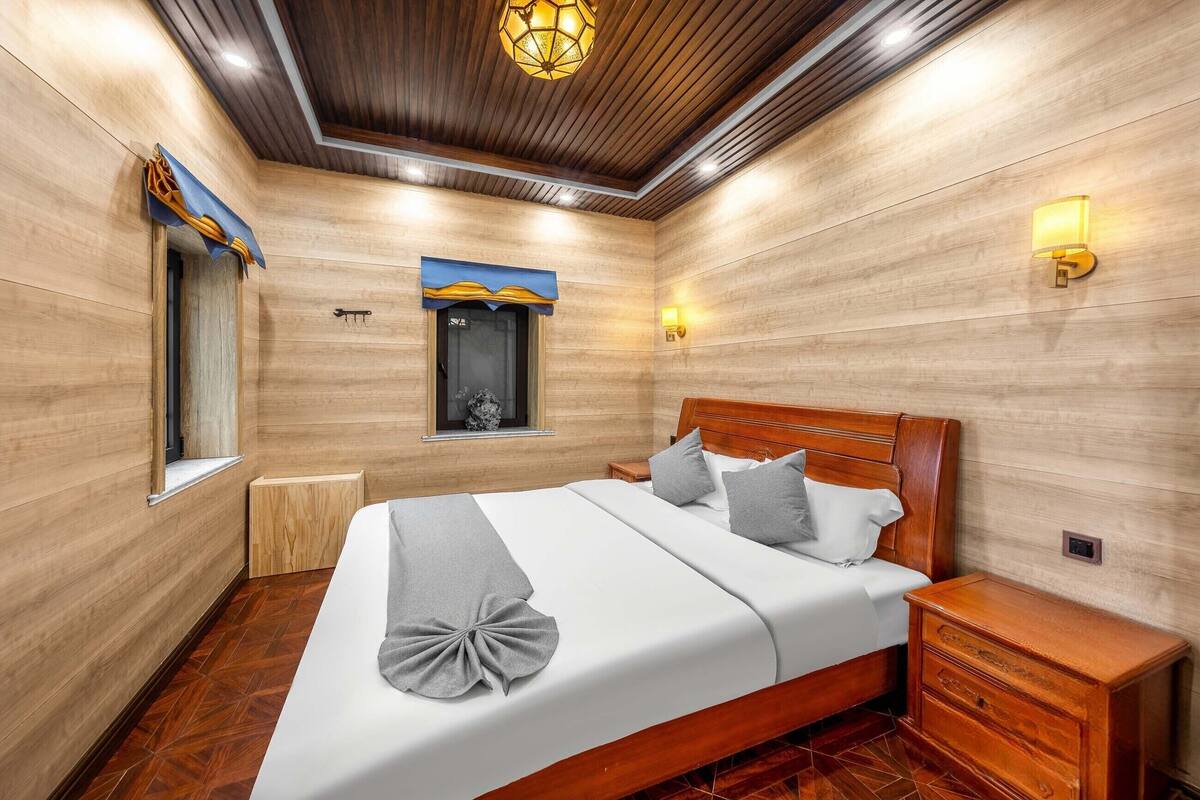 A hotel room with a wooden ceiling, a large bed with a headboard, two bedside tables, and a window with a curtain.