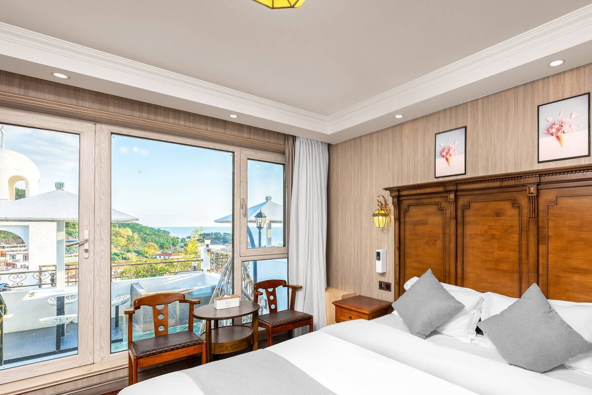 A hotel room with a large bed, a wooden headboard, a desk with a chair, and a view of the sea through large windows.
