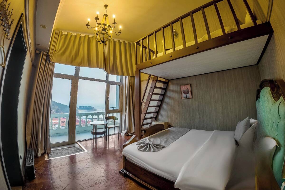 A bedroom with a bunk bed, a chandelier, a view of the sea, and a small table with chairs.