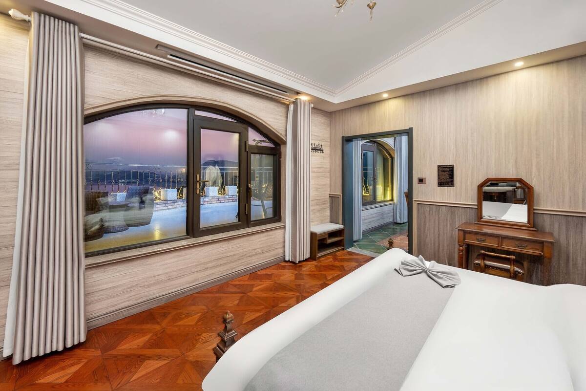 A hotel room with a large arched window, a wooden floor, a bed, a vanity with a mirror, and a bench.