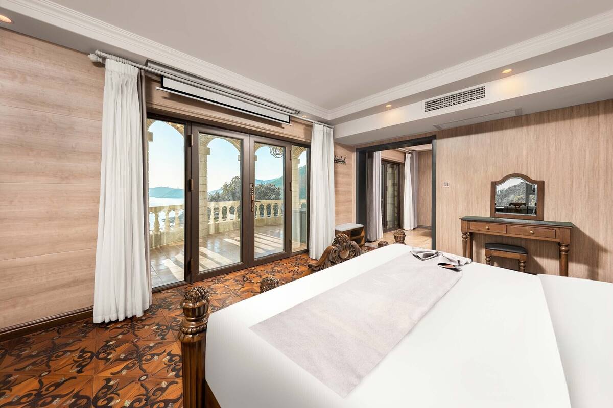 A hotel room with a large bed, a wooden desk, a mirror, and a view of the outdoors through a sliding glass door.