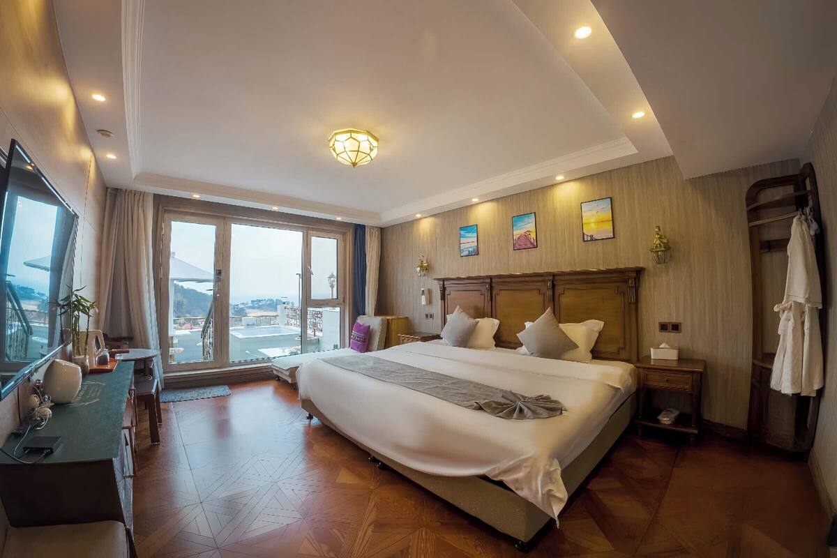 A spacious hotel room with a large bed, a desk with a chair, a television, and a view of the outdoors.