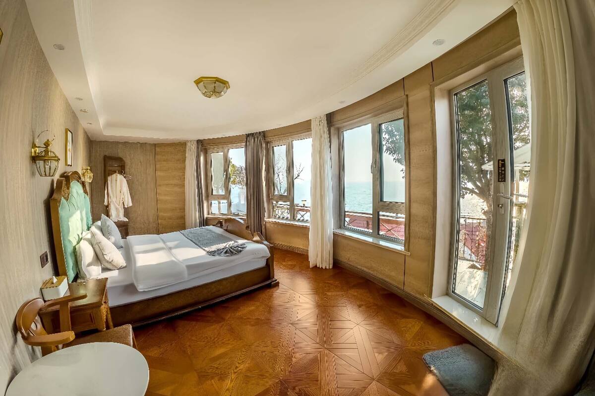 A spacious bedroom with a large bed, wooden flooring, and large windows offering a view of the sea.