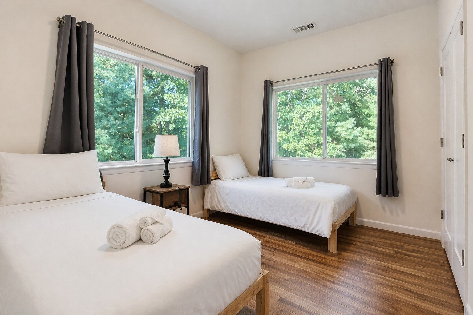 A bedroom with two beds, a nightstand with a lamp, and large windows with curtains.