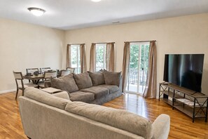 A living room with a sectional sofa, a dining table with chairs, a flat-screen TV on a stand, and large windows with curtains.