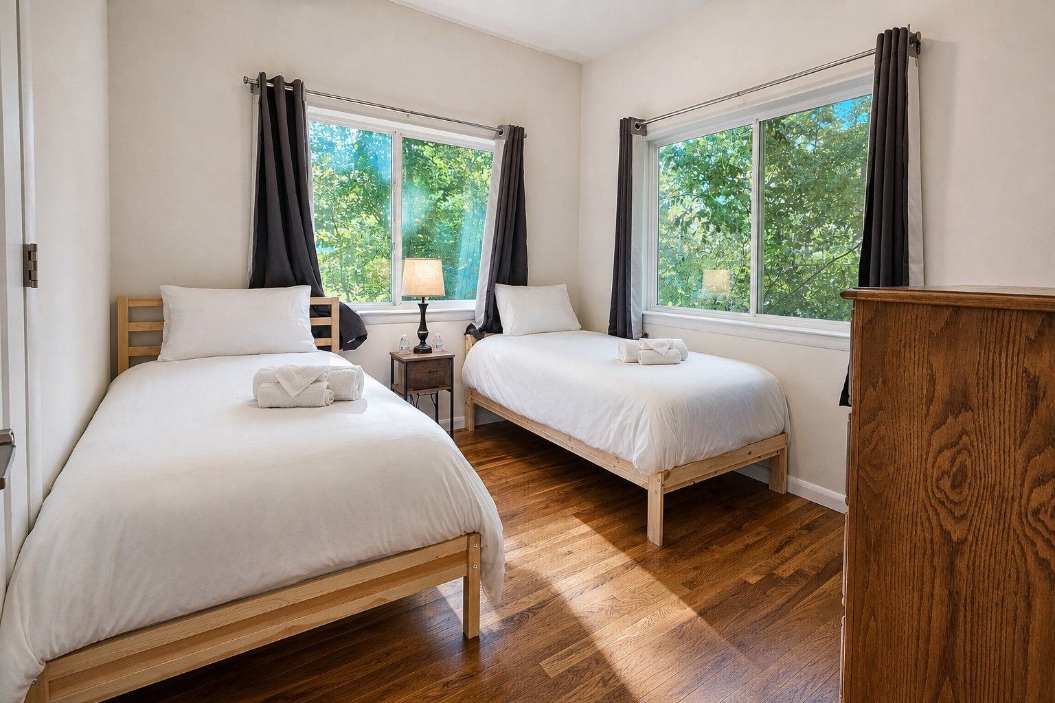 A hotel room with two single beds, wooden flooring, a nightstand with a lamp, and a view of greenery through the windows.