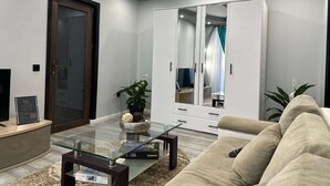 A modern living room with a glass coffee table, a beige sofa, a white wardrobe, and a large mirror. - G&A Luxe Apartment Central (Târgu Jiu)