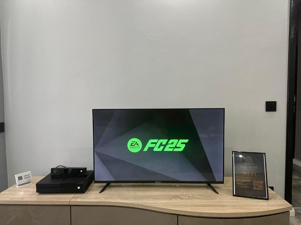 Un televisor de pantalla plana sobre un escritorio de madera con una consola de videojuegos y un cartel enmarcado.