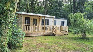 Une maison mobile dotée d’une terrasse en bois et d’un porche couvert, entourée d’une zone boisée.