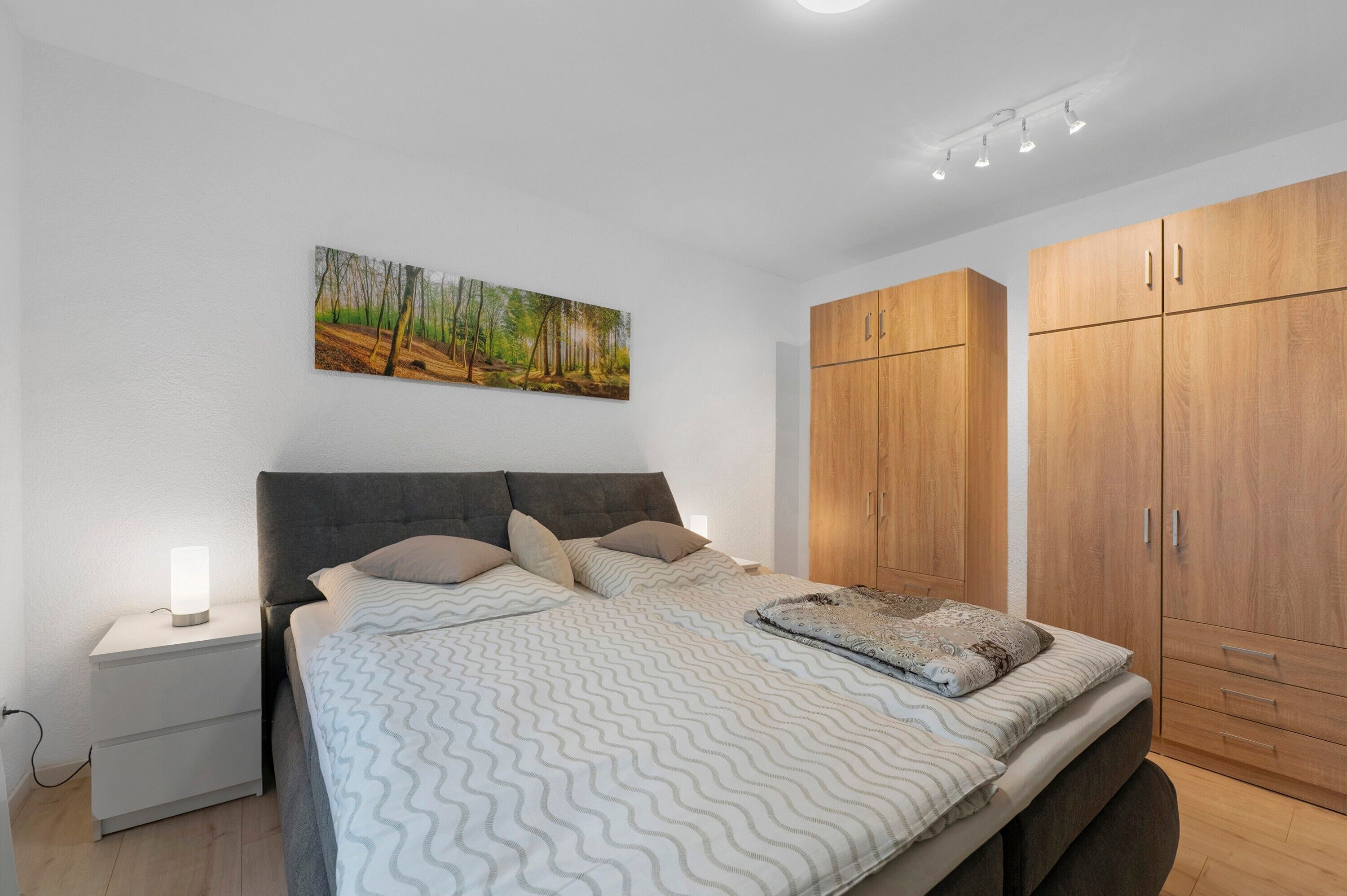 A bedroom with a bed, nightstand, wooden wardrobe, and a forest-themed wall art.