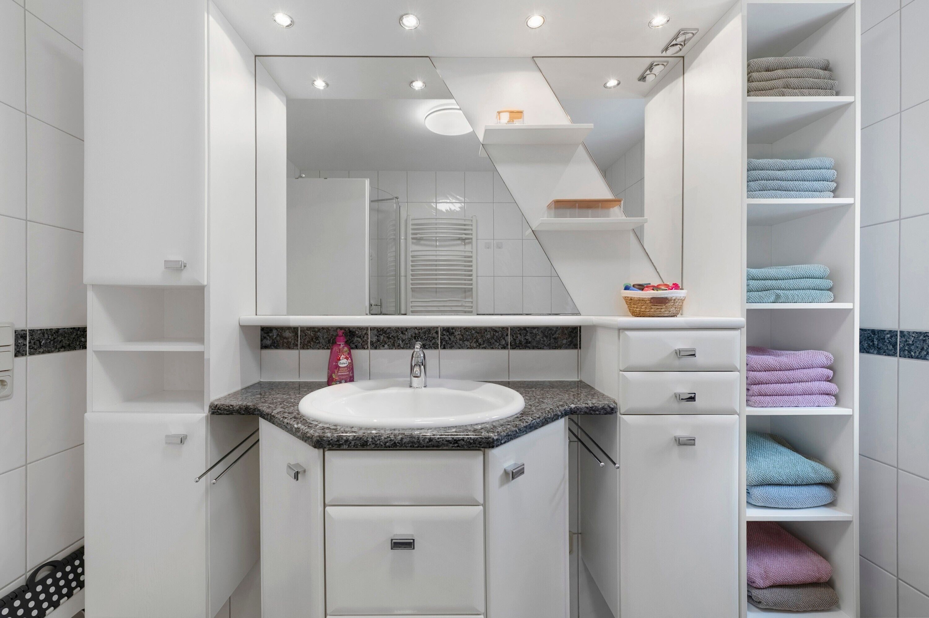 Ein modernes Badezimmer mit einem weißen Waschbecken, einer Arbeitsplatte aus Granit und einem großen Spiegel. Rechts befinden sich Regale mit gefalteten Handtüchern und ein Korb.