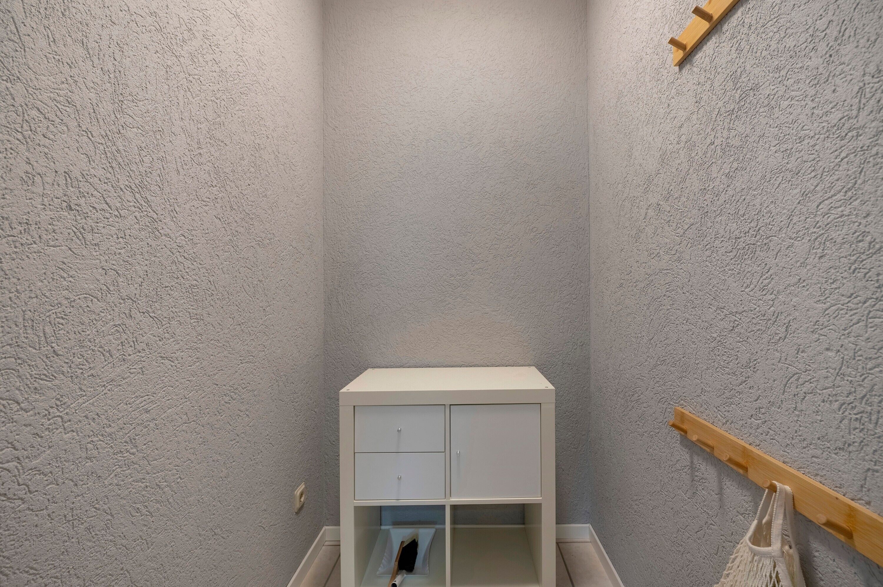 Ein kleiner, weißer Schrank mit zwei Fächern und einem Holzregal darunter, an einer strukturierten Wand.