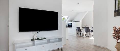 Una sala de estar moderna con televisor montado, mueble de entretenimiento blanco y un comedor al fondo.